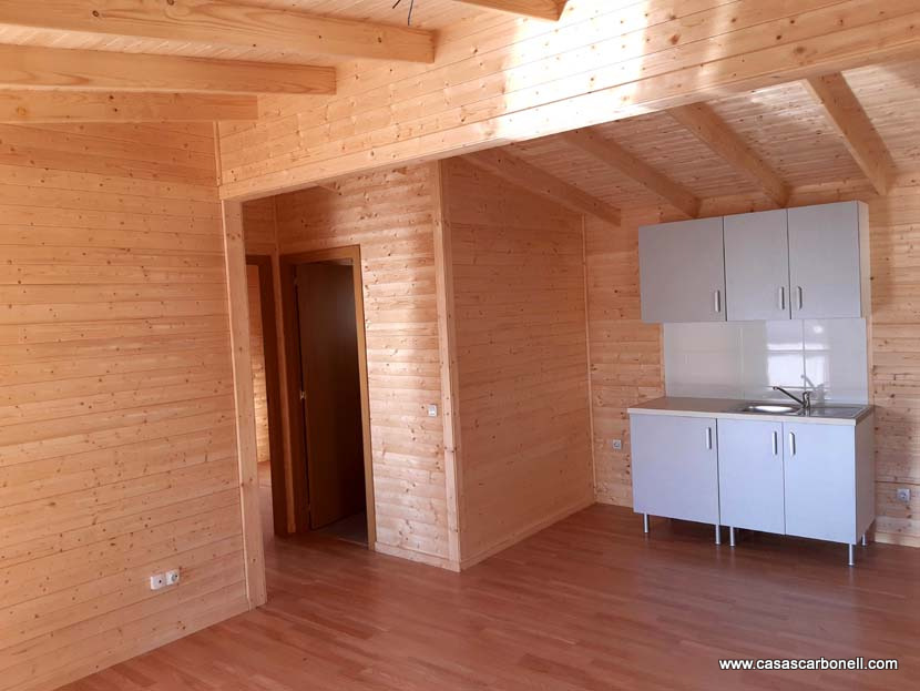 Interior de una casa de madera, con cocina integrada y techo con vigas.