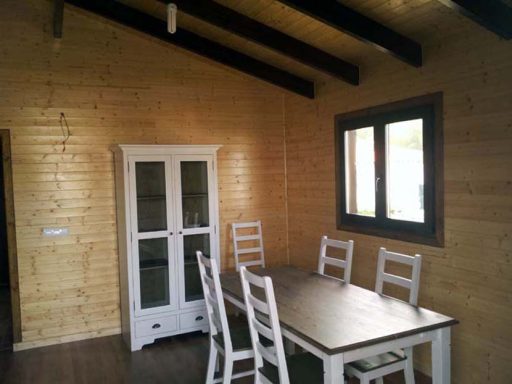 Interior de una cabaña con mesa, sillas blancas y paredes de madera clara.