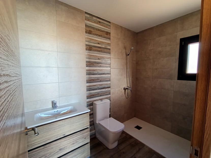 Baño moderno con ducha, lavabo, inodoro y azulejos en tonos beige y madera.